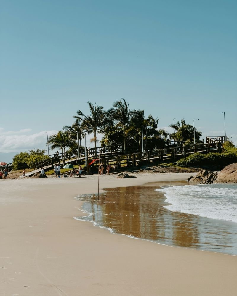 Passarela de madeira terceira pedra sobre a areia da praia em Itapoá, cercada por palmeiras e vegetação local, com mar calmo ao fundo e céu azul claro.